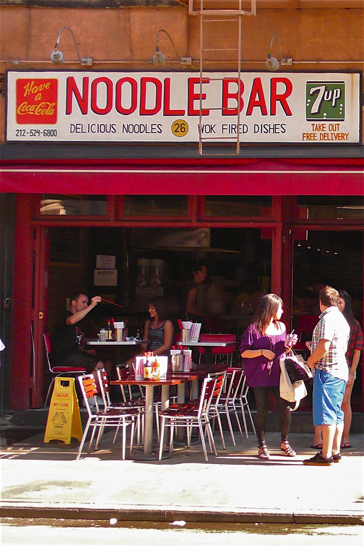 people are standing in front of a noodle bar with red awnings and tables outside