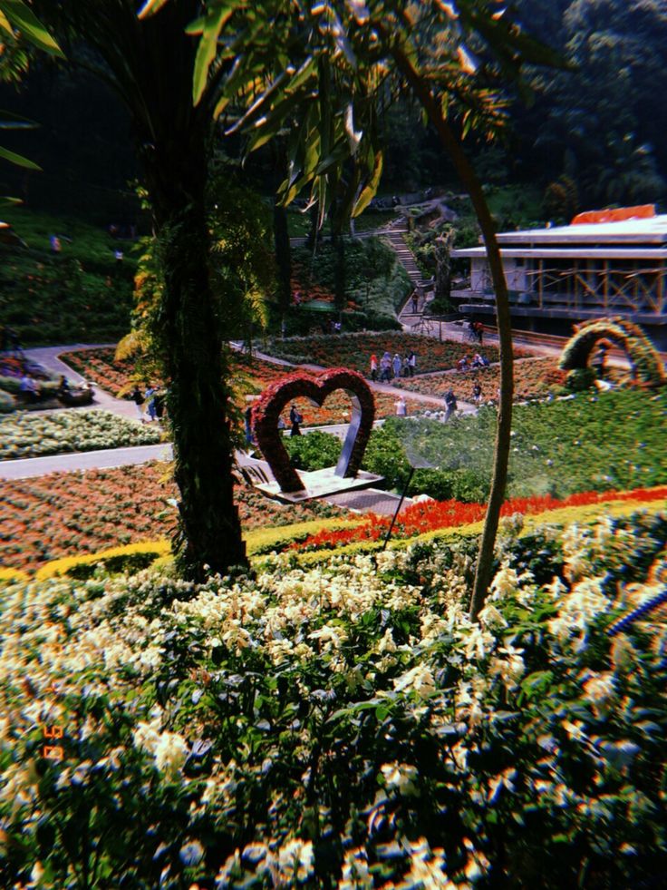 a heart shaped sculpture in the middle of a flower garden with trees and flowers around it