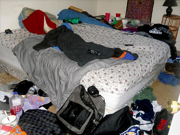 an unmade bed with lots of clothes on it and a bag next to it