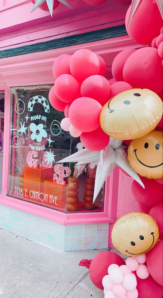 some balloons are hanging on the side of a pink store front window and it is decorated with smiley face balloons