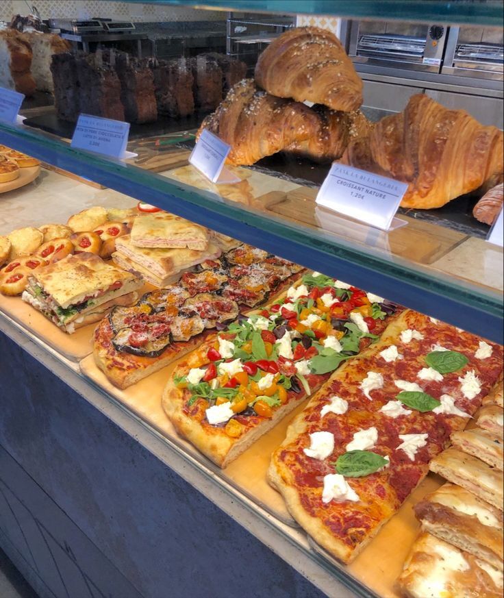 many different types of pizzas on display in a store window with croissants and bread