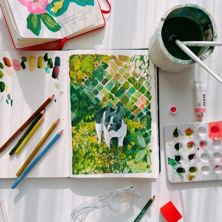 an open book with watercolors, paint and pencils next to it on a table