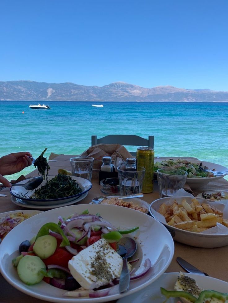 a table with plates and bowls of food on it near the water's edge