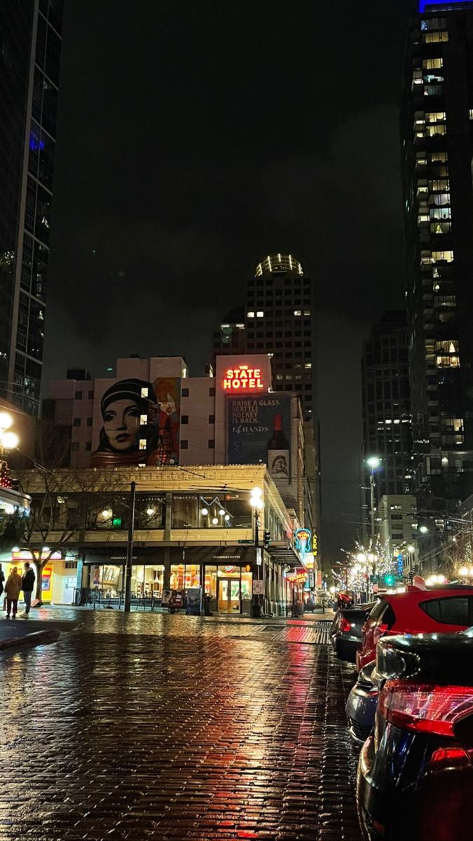 cars are parked on the street in front of some tall buildings at night with people walking by