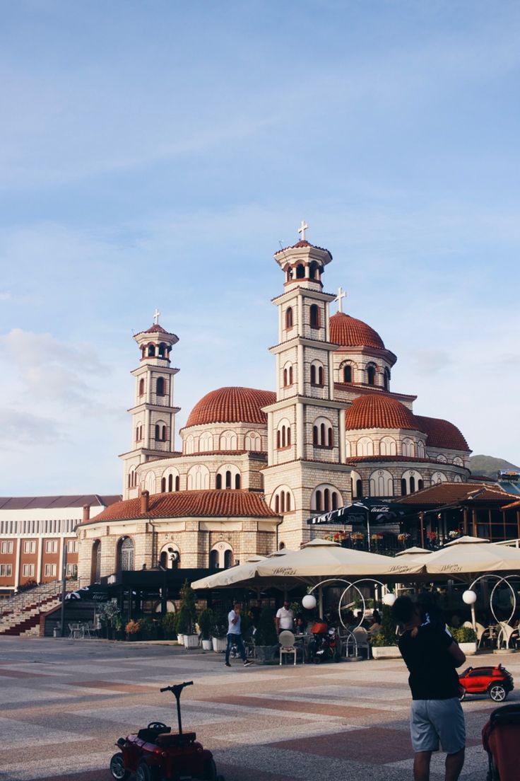 two people standing in front of a large building with red roof tops and white trim