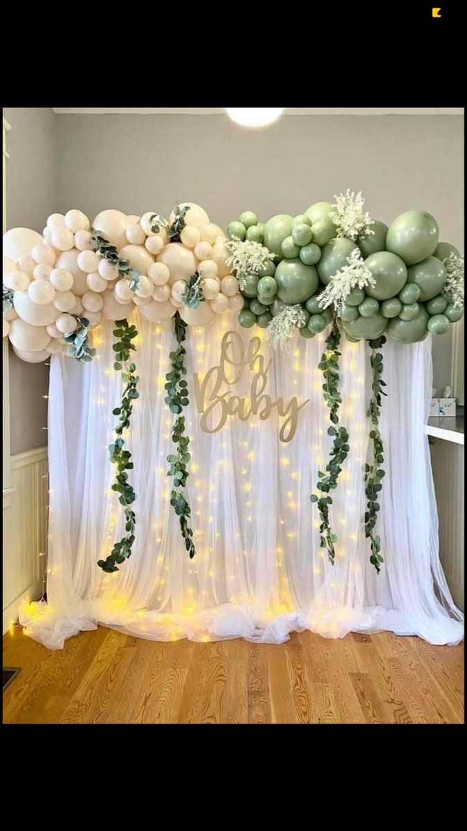 balloons and greenery decorate the backdrop for a baby shower with white sheer curtains, green garlands and lights