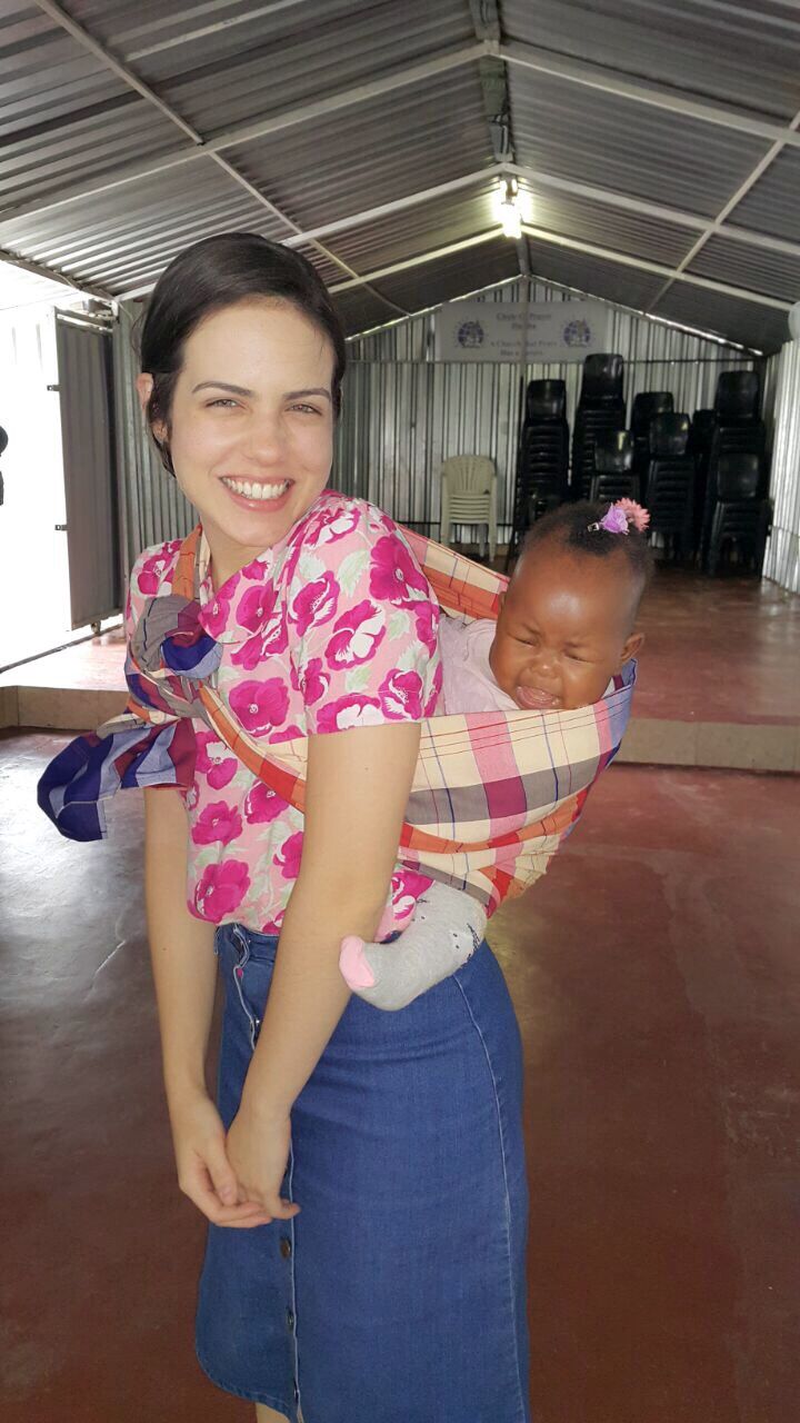 a woman carrying a baby in a pink and blue wrap on her back while smiling at the camera