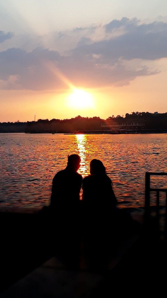 two people sitting next to each other on a pier watching the sun go down over water