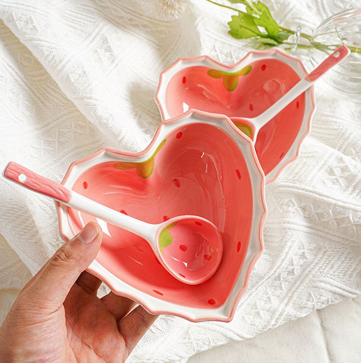 two pink heart shaped bowls with spoons in them on a white surface next to a flower