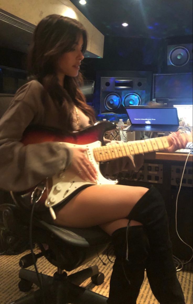 a woman sitting in front of a laptop computer while playing an electric guitar on top of a desk