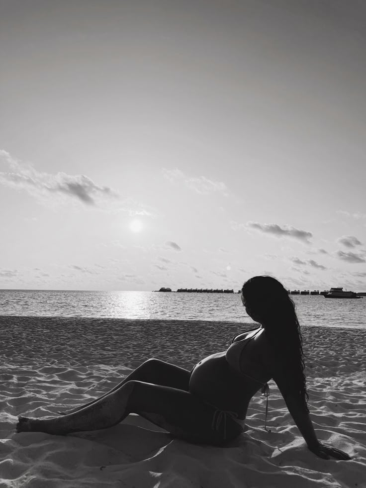 a woman sitting on top of a sandy beach next to the ocean in black and white