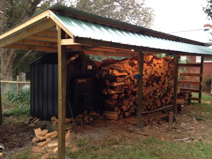 a pile of logs sitting under a metal roof next to a shed with a green tin roof