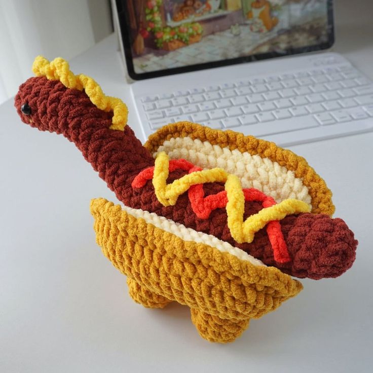 a crocheted hot dog on a bun with mustard and ketchup in front of a laptop computer