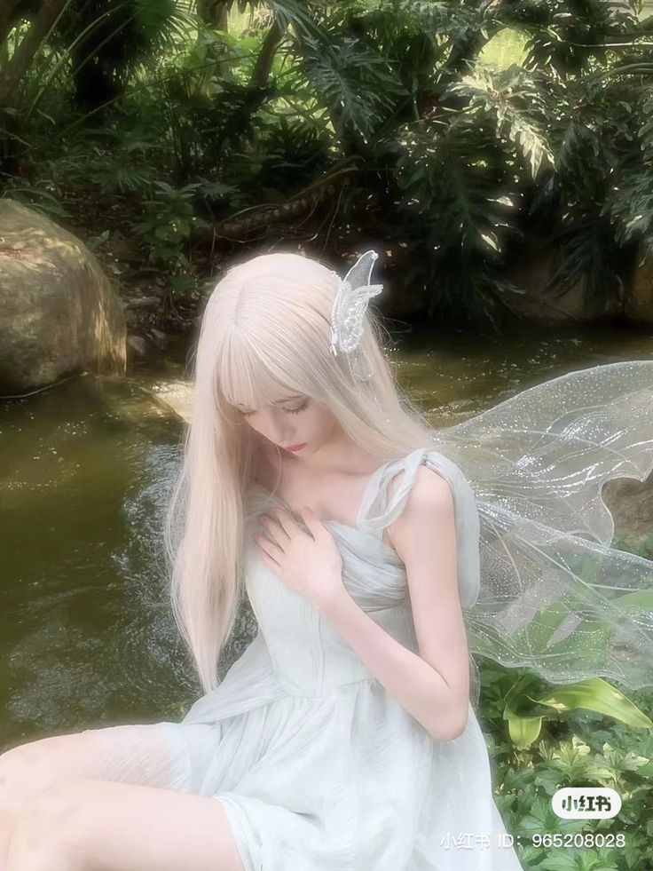 a woman with long blonde hair sitting on the ground next to some water and plants