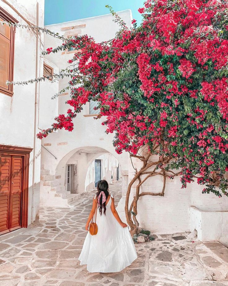 a woman in a white dress is walking down an alley way with red flowers on the tree