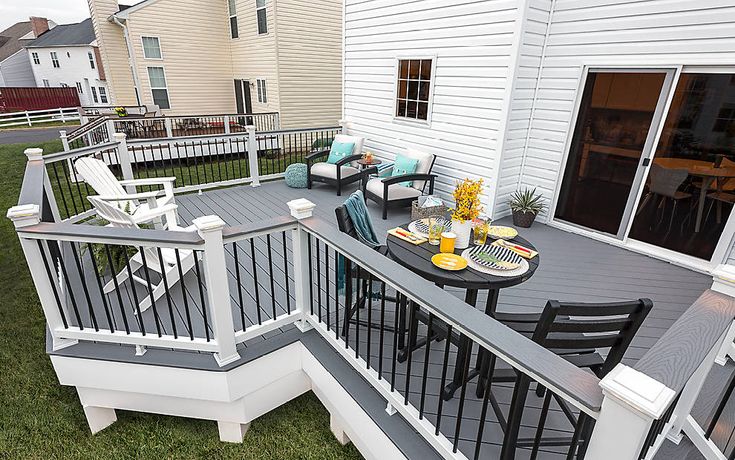 an outdoor deck with chairs and table next to a white house on the grass in front of it