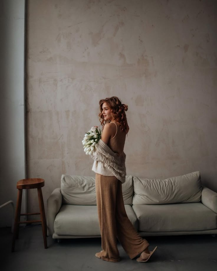 a woman standing in front of a couch holding a bouquet of flowers and looking at the wall