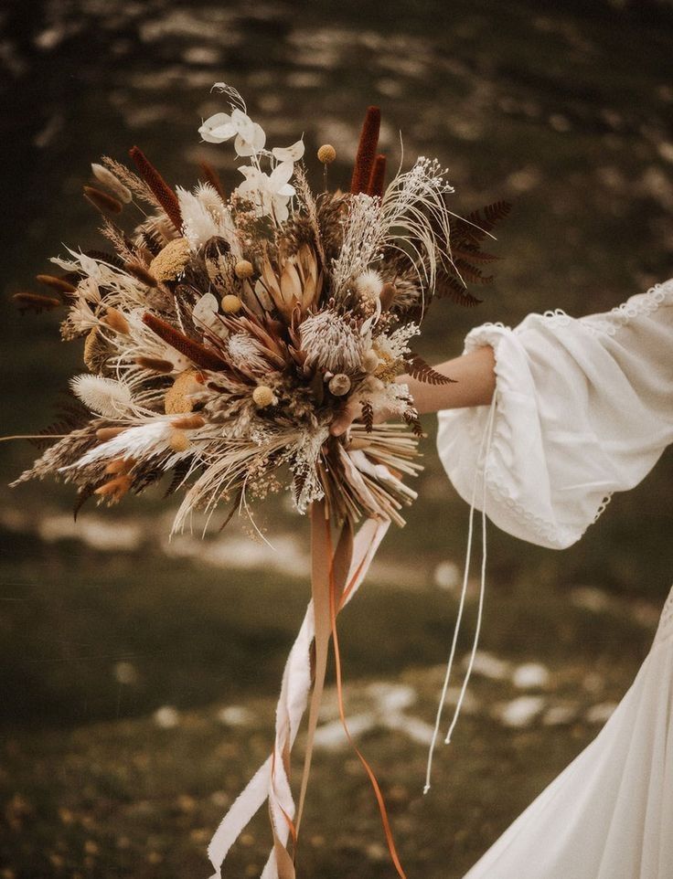 a bride holding a bouquet of dried flowers and feathers in her hand with the wind blowing