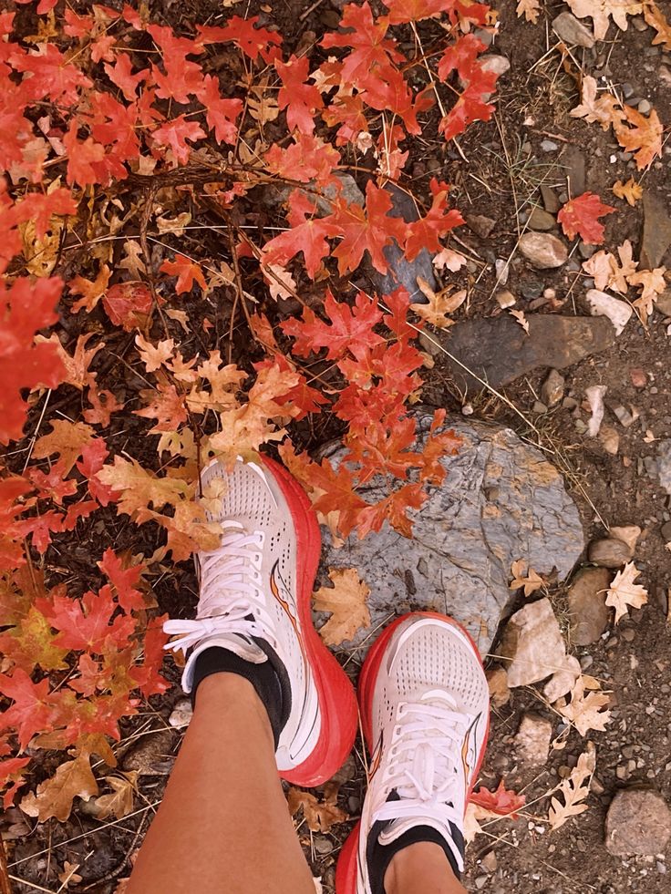 a person's feet with red and white tennis shoes in front of autumn leaves