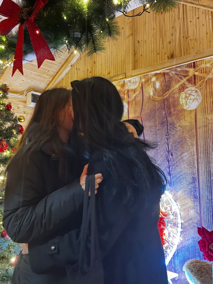 two women standing next to each other in front of a christmas tree with lights and decorations