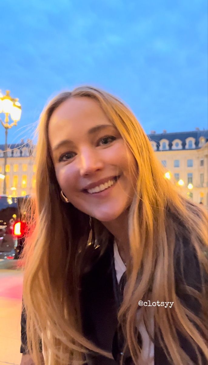 a woman with long blonde hair smiling at the camera in front of a large building