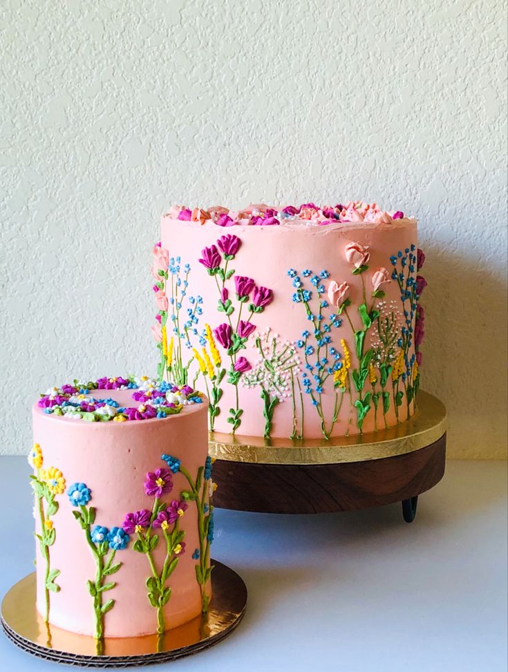 two cakes decorated with pink frosting and colorful flowers on wooden stands next to each other