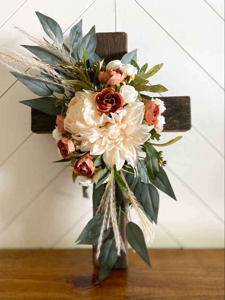 a cross decorated with flowers and greenery on a wooden table in front of a wall