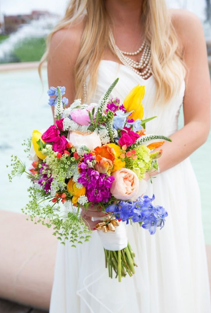 a woman in a white dress holding a bouquet of colorful flowers and greenery near a fountain