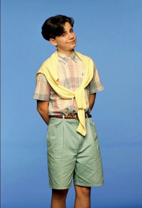 a young man wearing shorts and a yellow scarf over his shoulders, standing in front of a blue background