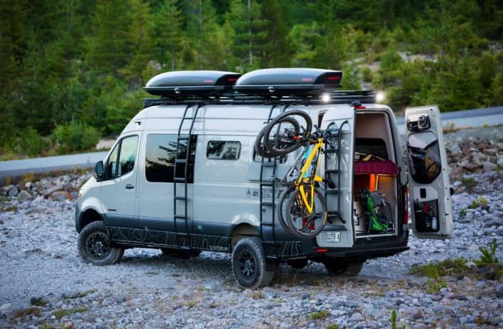 a van with bikes on the back parked in gravel area next to trees and rocks