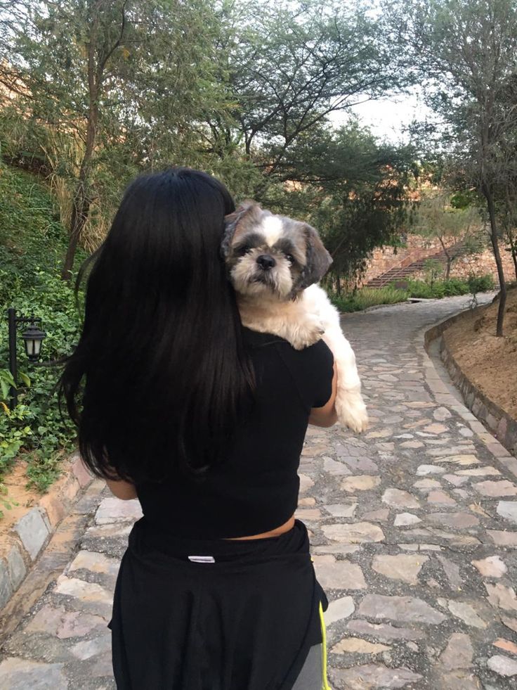 a woman holding a small dog in her arms on a cobblestone path with trees and bushes behind her