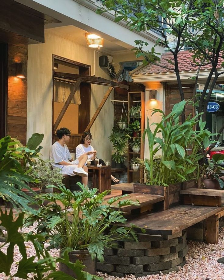 two people sitting at a table in front of a house with plants and potted trees