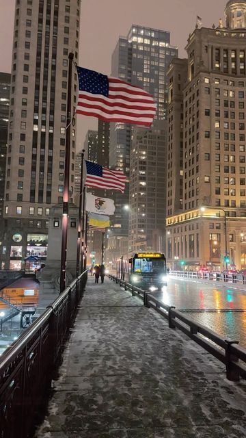 an american flag flying in the wind on a city street at night with tall buildings