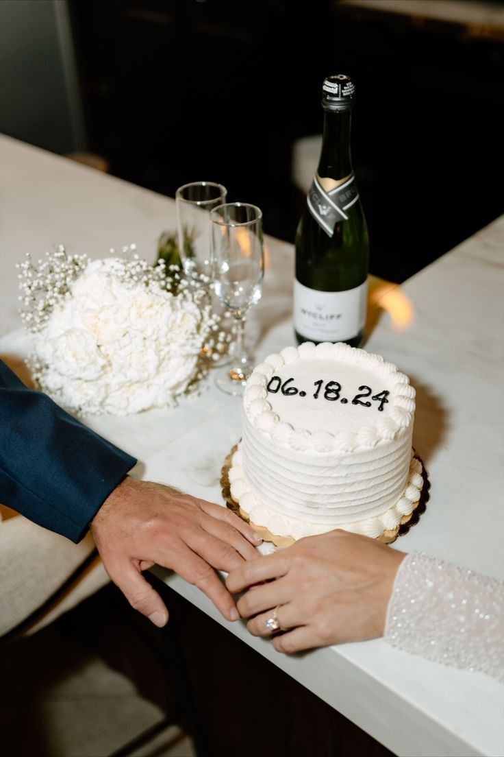 two people holding hands over a cake on a table next to wine bottles and glasses