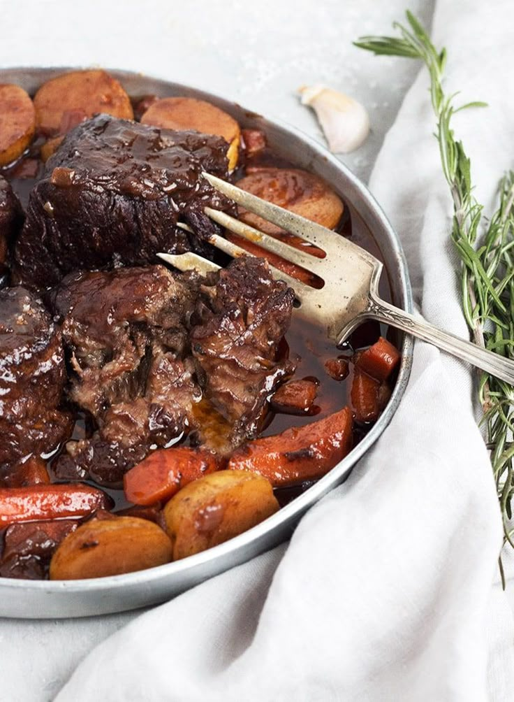 a pot filled with meat and vegetables on top of a white cloth next to a fork