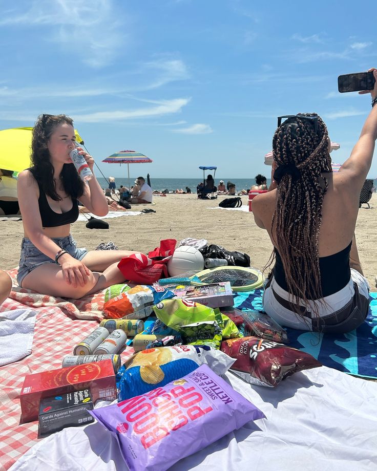 two women are sitting on the beach with snacks