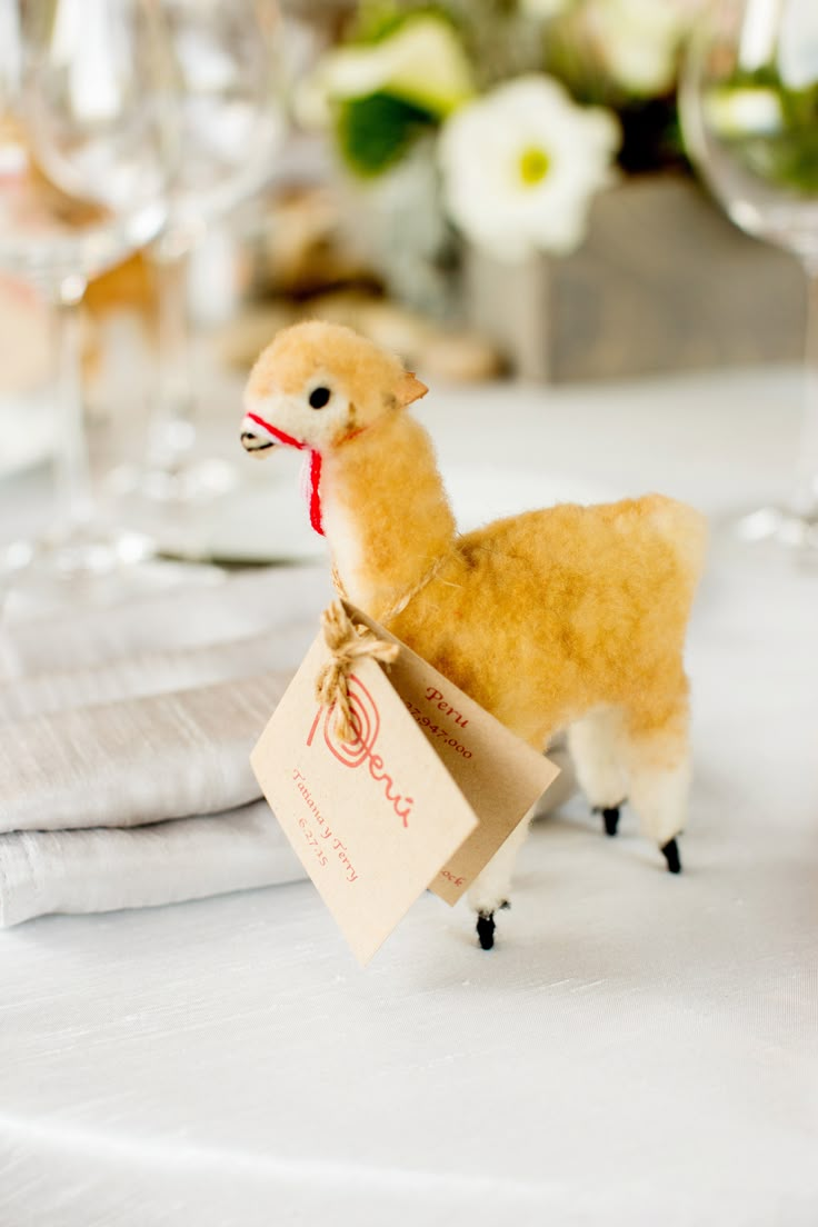 a small stuffed llama holding a tag on top of a white table cloth with wine glasses and flowers in the background