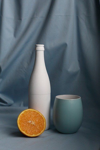 an orange sitting next to a white vase on a gray background with a blue cup