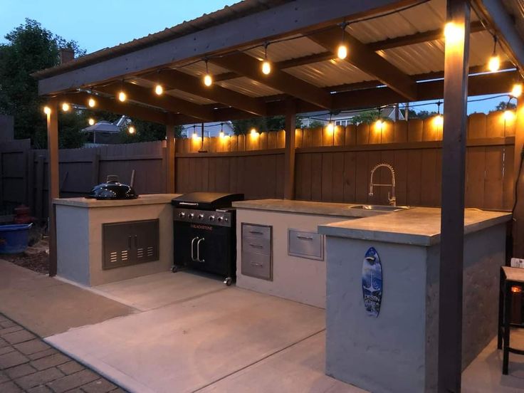 an outdoor kitchen is lit up at night with string lights on the roof and grill area