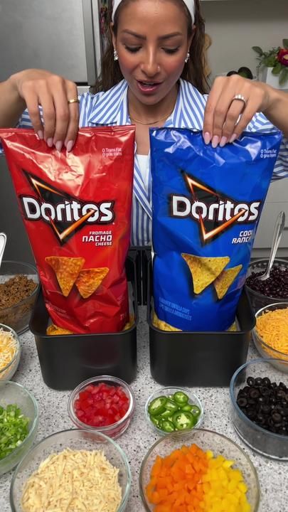 a woman holding two bags of doritos next to bowls of beans and vegetables