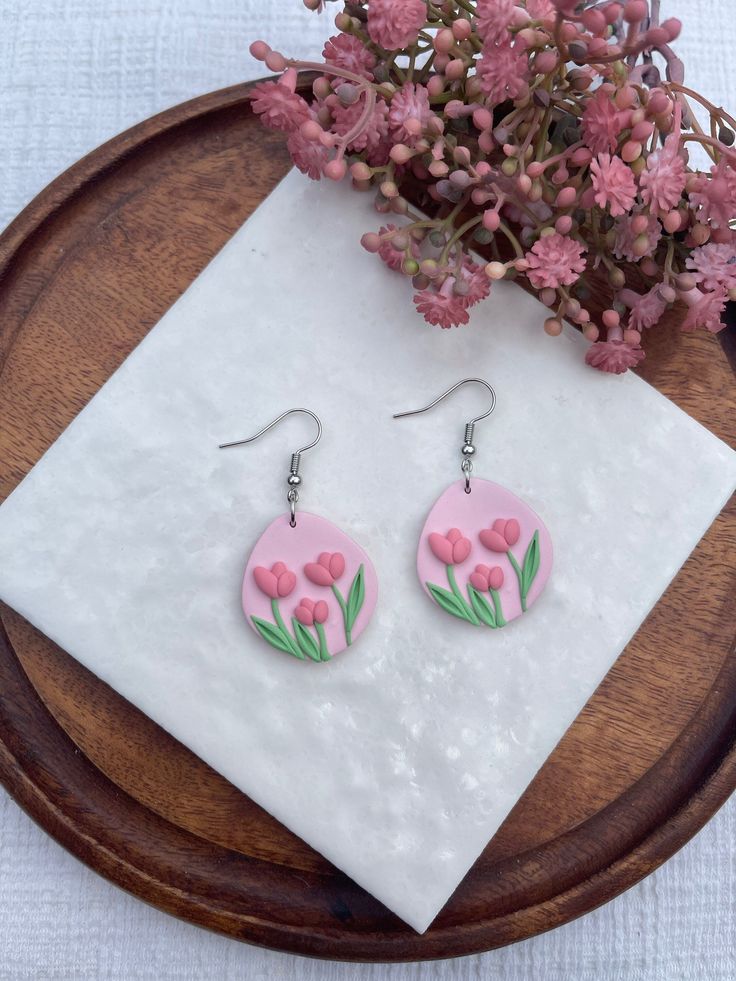 a pair of pink flower earrings sitting on top of a wooden plate next to flowers