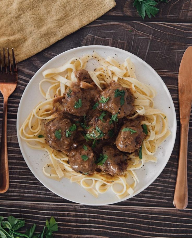 a white plate topped with meatballs and pasta next to silverware on top of a wooden table