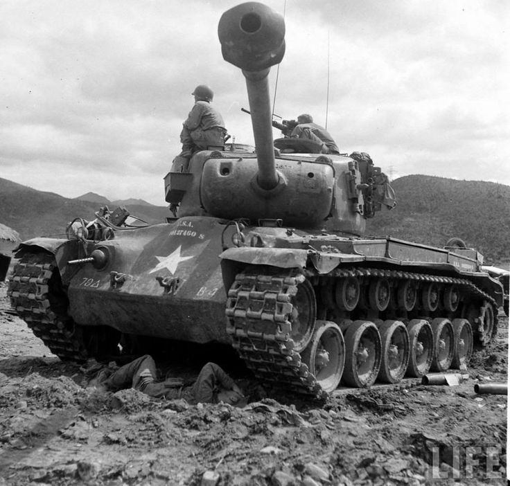 an old tank with two men on top of it in the mud and mountains behind it