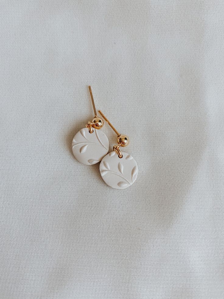 two white earrings are hanging from gold earwires on a white surface with light colored fabric