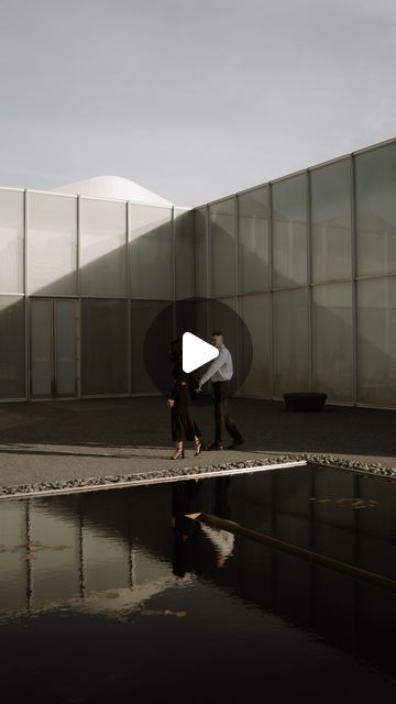 two people are walking in front of a building with glass walls and reflecting water on the ground