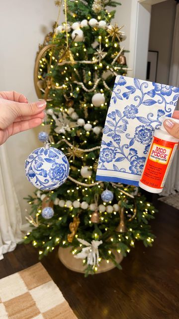 two hands holding up a blue and white christmas ornament next to a decorated christmas tree