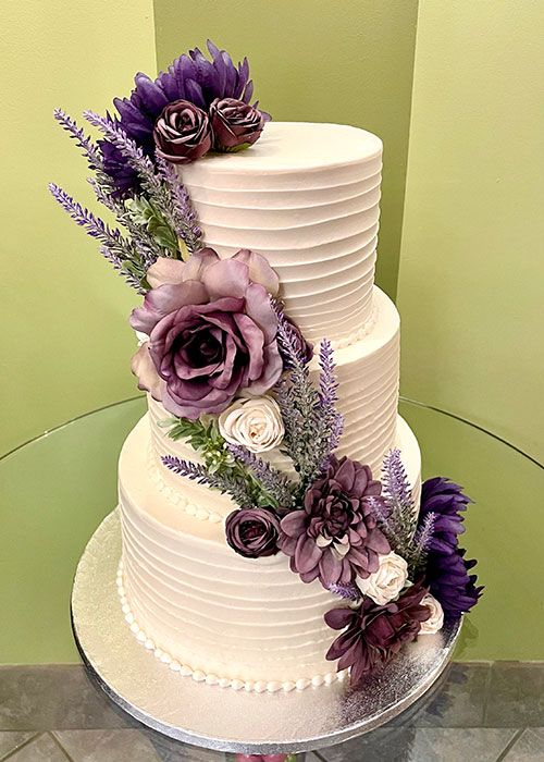 a three tiered white cake with purple flowers on the top and bottom, sitting on a glass table