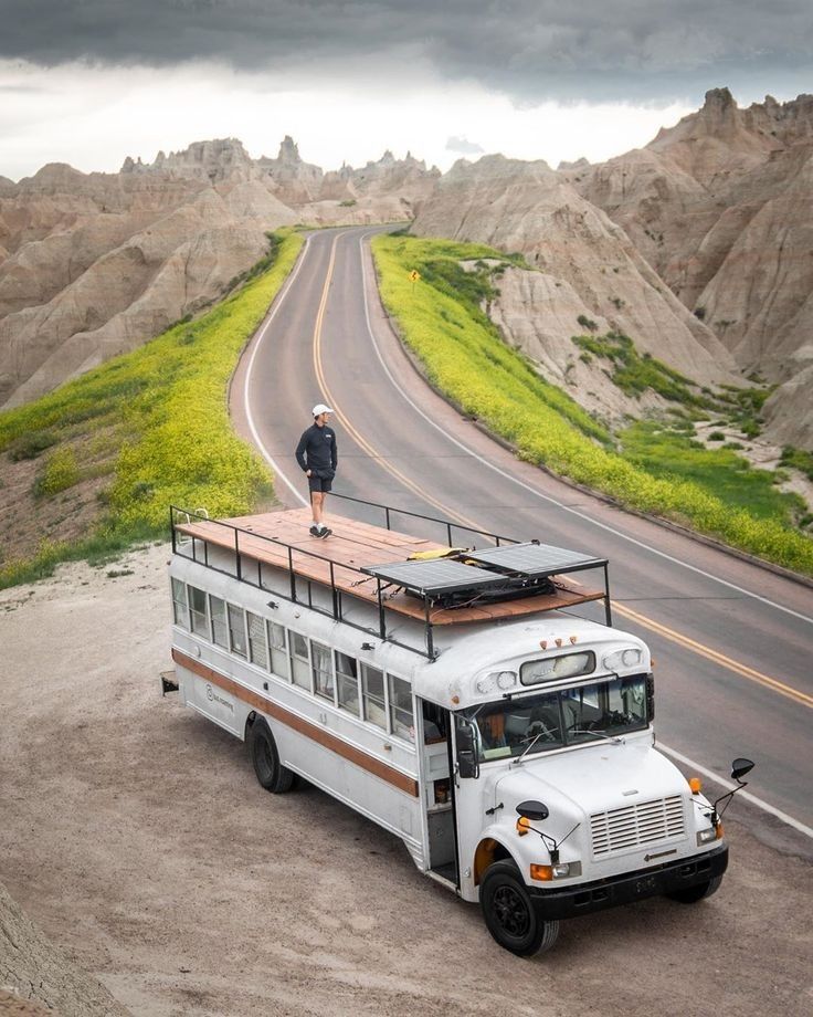 a man standing on top of a bus in the middle of an empty road with mountains behind it