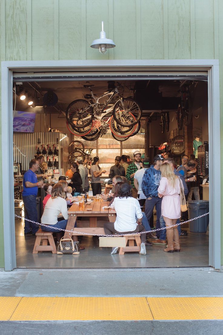 a group of people sitting at a table in front of a bike shop door with bicycles hanging from the ceiling