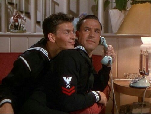 two men sitting next to each other on a couch talking on the phone and holding something in their hands